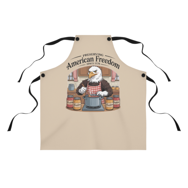 1045375229664899763_2048.webp Apron with a cartoon graphic of an eagle wearing a red and white checkered apron, stirring a pot in a kitchen scene with jars labeled "Freedom".