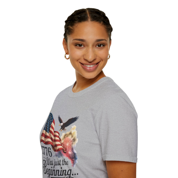 A smiling woman wearing a gray t-shirt with a patriotic design including an American flag, an eagle, fireworks, and the text '1776 Was just the Beginning...'