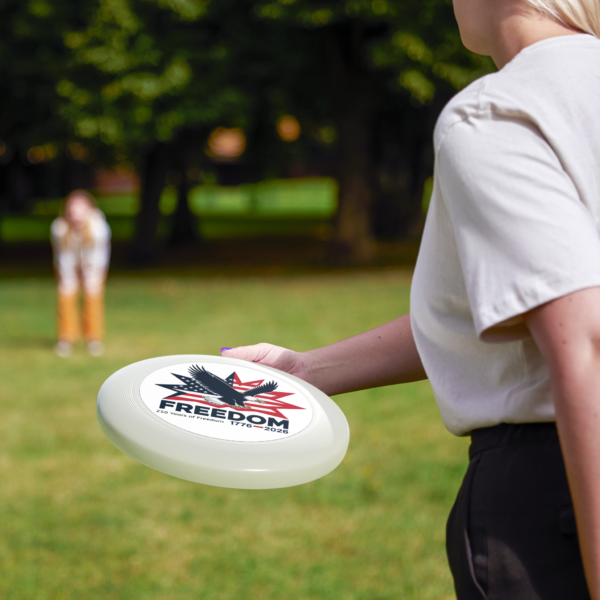 13749270903011980509_2048.webp A woman is holding a white frisbee with an American eagle and the word 'Freedom' emblazoned on it, against a blurred green background.