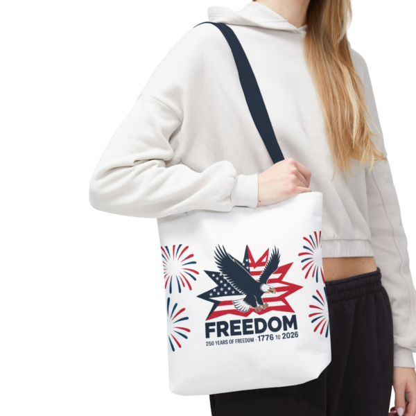 A woman holds a white tote bag with a patriotic design featuring an eagle, stars, stripes, and the text 'Freedom 250 years of freedom - 1776 to 2026'.