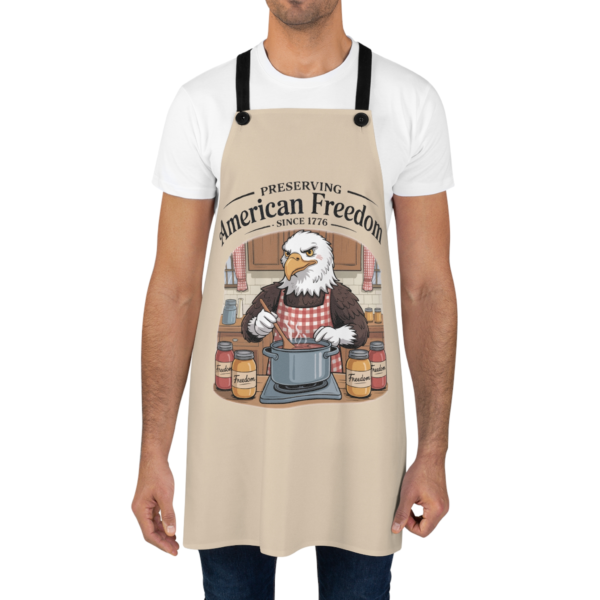 18408020081684021204_2048.webp A person wearing a beige apron with a cartoon eagle stirring a pot in a kitchen scene and the words 'Preserving American Freedom Since 1776'.