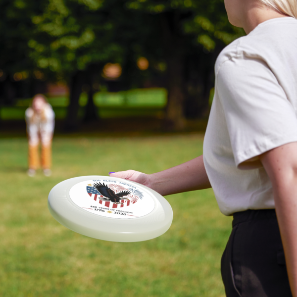 2965755545399903839_2048.webp A woman holds a white frisbee featuring an American flag, an eagle, and fireworks with the text 'God Bless America' and '250 Years of Freedom 1776-2026'.
