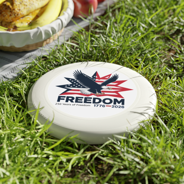 5904021158906312323_2048.webp White frisbee lying in grass, featuring a patriotic design of an eagle in front of a starburst of American flag stripes, and the word 'FREEDOM' above '250 Years of Freedom 1776-2026'.