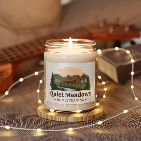 5945444513485016760_2048.webp A lit 'Quiet Meadows Homesteading' candle in a glass jar, featuring a label with a homestead scene.